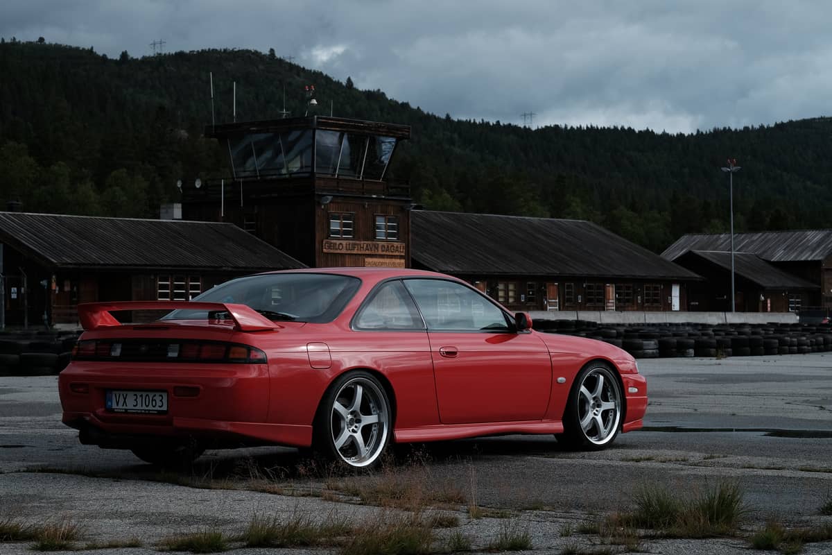 Rød Nissan Silvia parkert på asfalten ved Geilo Lufthavn Dagali, med mørke tømmerbygg, dekkstabler og skogkledde åser under lavt skydekke.